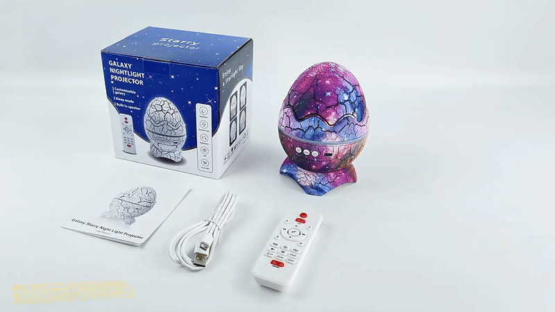 Night Sky Projector shaped like a cracked dino egg with galaxy design, remote control, USB cable, and packaging box for cosmic room lighting and Bluetooth audio.