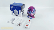 Night Sky Projector shaped like a cracked dino egg with galaxy design, remote control, USB cable, and packaging box for cosmic room lighting and Bluetooth audio.